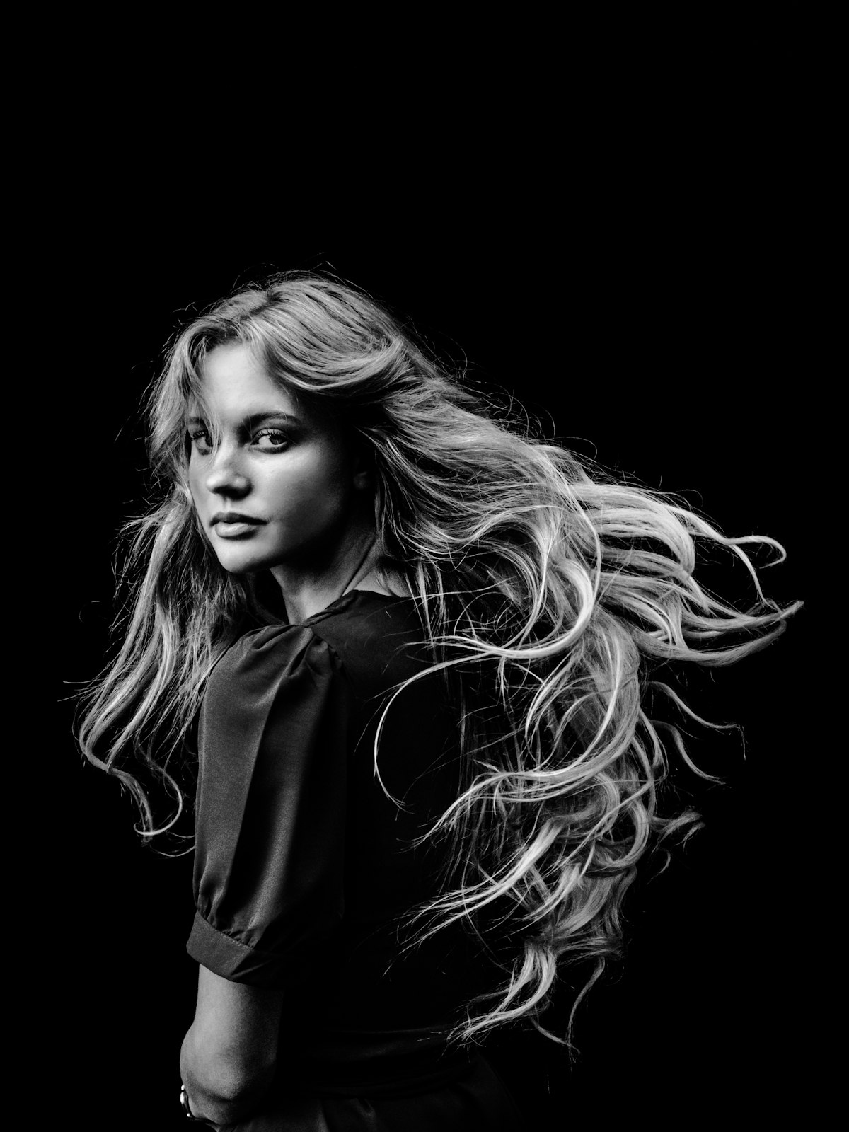 Black-and-white studio portrait: long hair caught mid-movement against a dark ground.