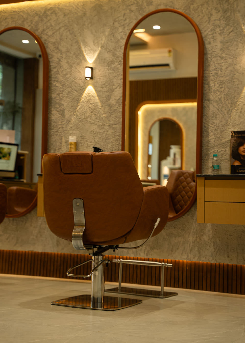A tan leather styling chair standing between two arch-topped mirrors on a textured plaster wall, lit by a single wall lamp.