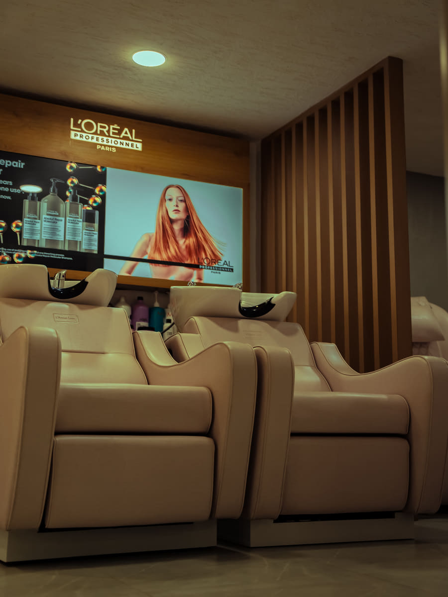 Two cream reclining basin chairs beneath a wall-mounted screen showing a hair campaign.