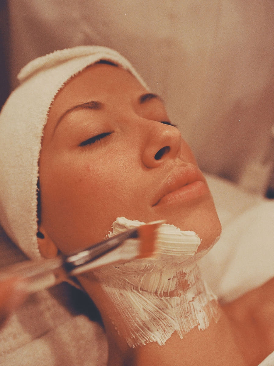 A treatment mask being brushed along a client's jaw and neck during a facial, her hair wrapped in a white band.