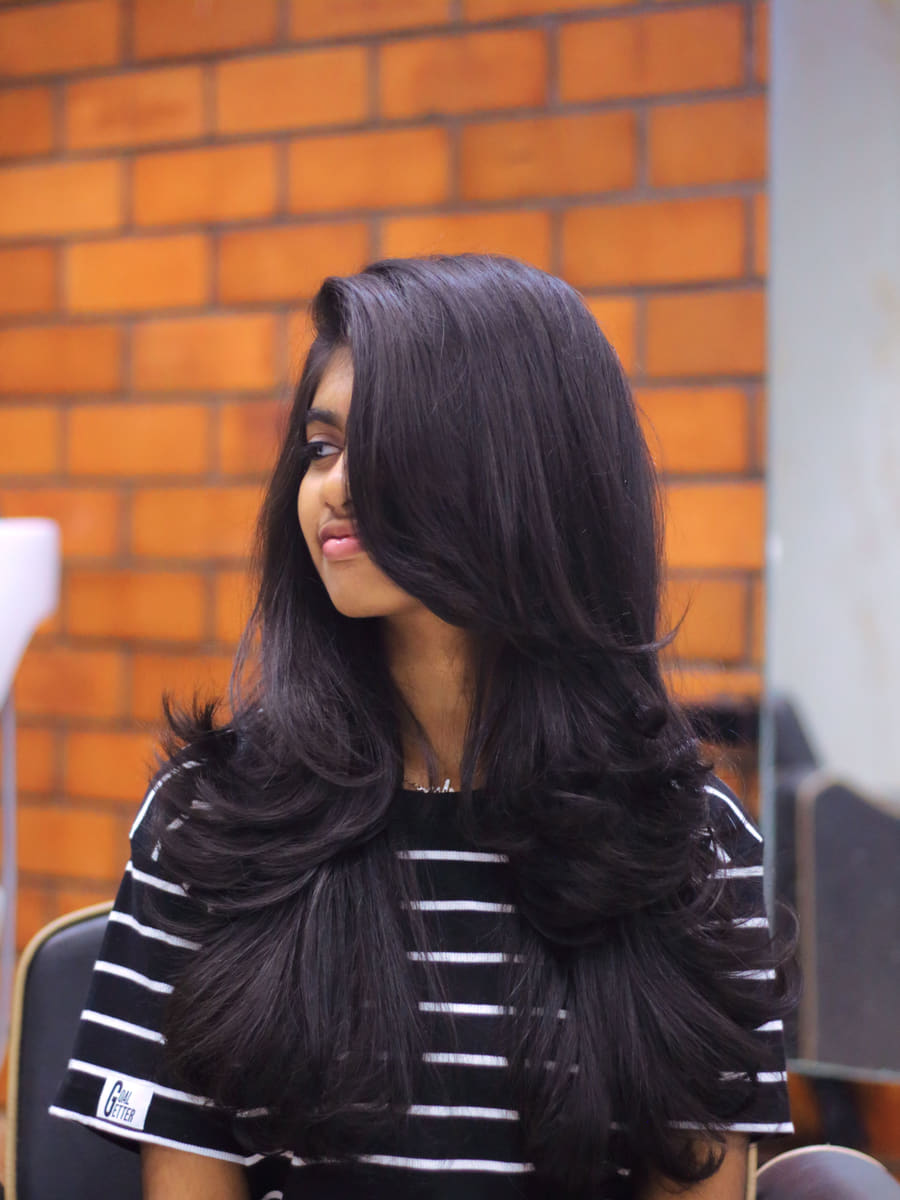 A client with long dark hair blow-dried into deep face-framing layers, photographed in profile against a brick wall.